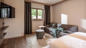 A modern hotel room with a flat-screen TV, a sofa, a small round table, and a window with curtains.