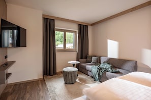 A modern hotel room with a flat-screen TV, a sofa, a small round table, and a window with curtains. - by franz - Alpine Apartments (Kirchberg in Tirol)