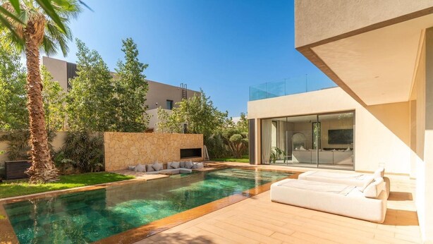 A modern outdoor pool area with a wooden deck, a stone wall, and a white outdoor seating area. - Palais Eden (Marrakech)