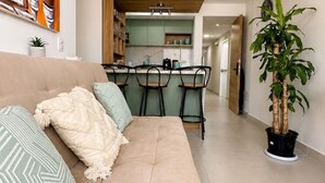 Een woonkamer met een bank, een eethoek met stoelen, een keuken op de achtergrond en een kamerplant.