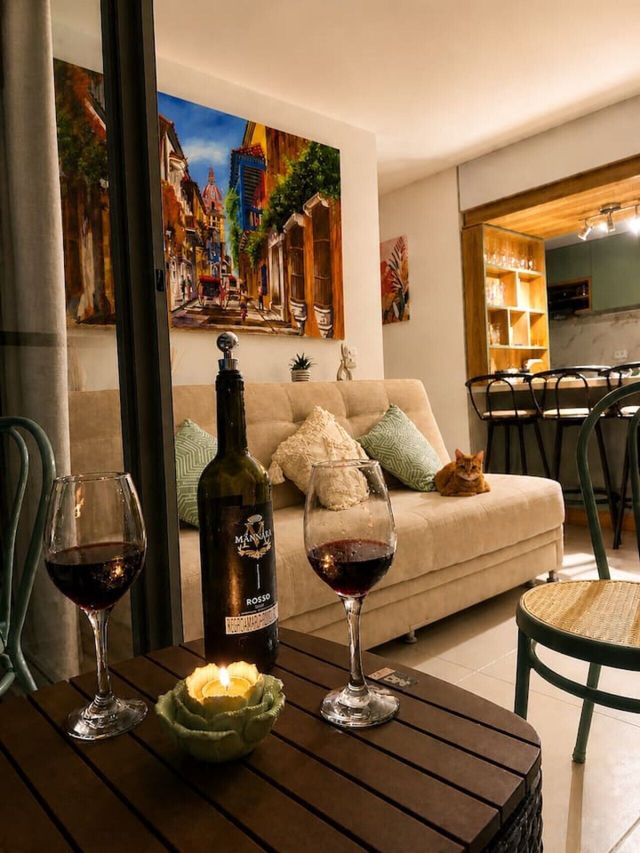 A living room with a sofa, a table with wine glasses and a bottle, and a painting of a street scene.