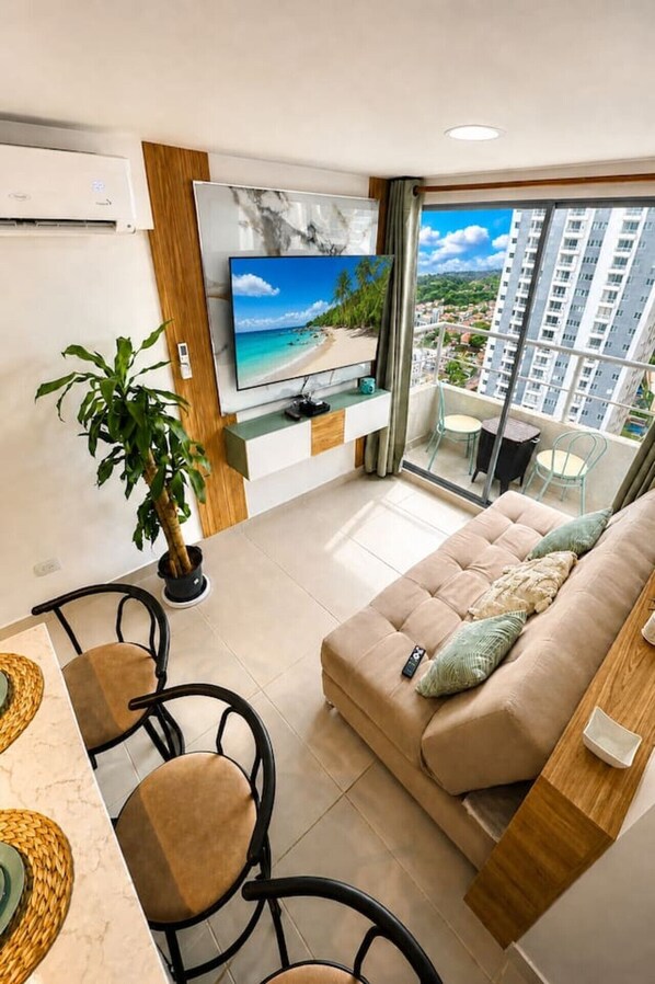 A modern living room with a flat-screen TV, a sofa, a dining area, and a balcony with a view of a beach and high-rise buildings. - Caribbean Deluxe • Pool, Views & WiFi (Cartagena)