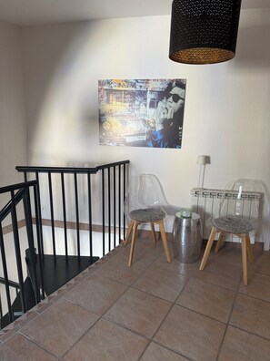 A staircase with a black railing, a clear plastic chair with a fluffy seat, a metal trash can, and a wall-mounted lamp. - Dream Home for Rent in Southern France (Soubès)