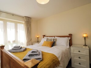 Ein ordentlich gemachtes Bett mit Kissen und Decke, ein Nachttisch mit Lampe sowie ein Fenster mit Vorhängen.
