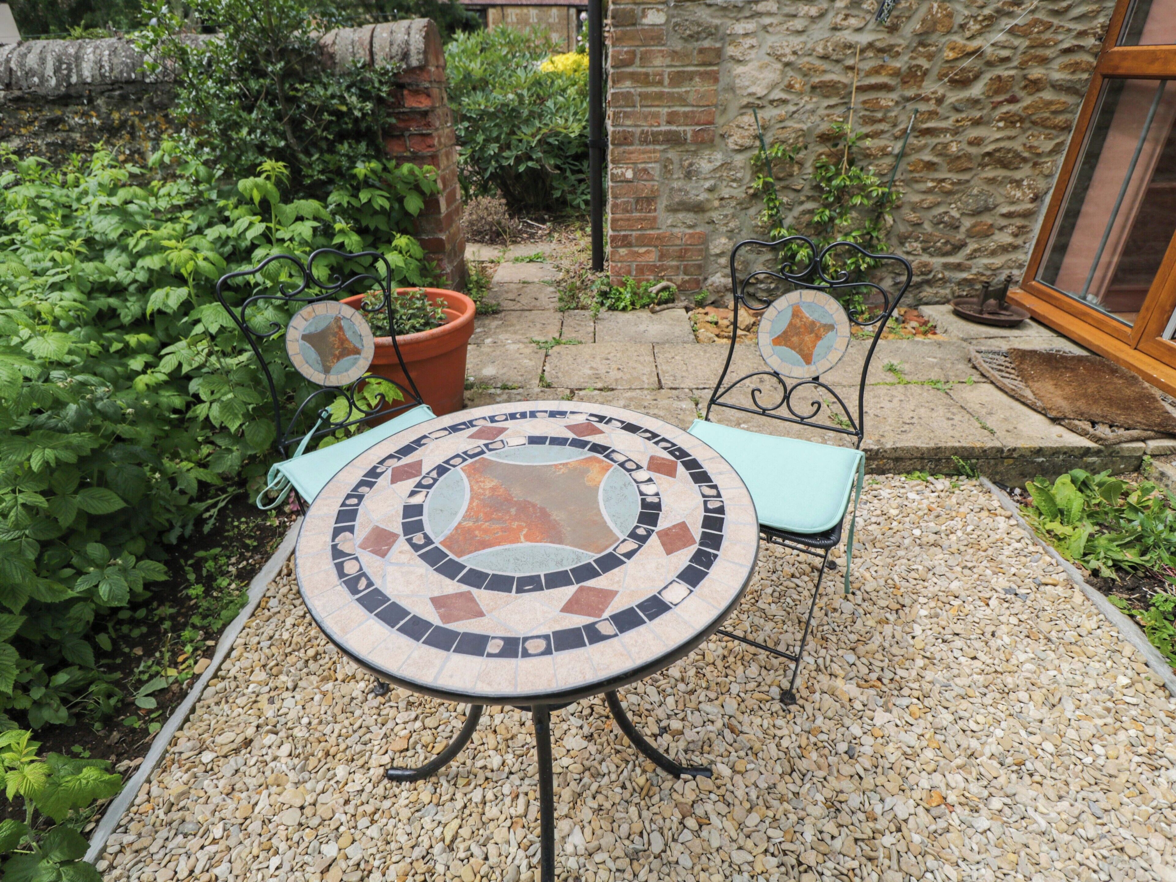 Een ronde tafel met mozaïektegels, twee stoelen, een bloempot en een stenen muur in een tuin.