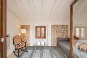 A room with a patterned floor, a wooden rocking chair, a lamp, a stone wall, a wooden door, and a gray sofa with pillows. - Holiday Home 'Rectoria 8' with Mountain View, Wi-Fi and Air Conditioning (Valldemossa)