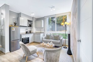 Une cuisine moderne avec des meubles gris, un four encastré et un réfrigérateur. On y trouve un canapé gris avec des coussins, une table basse et un lampadaire.