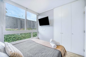 Ein Schlafzimmer mit Bett, einem an der Wand montierten Fernseher, einem Fenster mit Blick auf Grünflächen und einem Kleiderschrank.