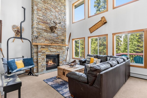 A living room with a stone fireplace, leather sofa, and large windows with a view of trees. - Summit County Escape | Private Hot Tub+ Deck (Breckenridge)