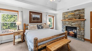 Ein Schlafzimmer mit Kamin aus Stein, einem Bett mit Holz-Kopfteil, einer Bank und einem Fenster mit Blick auf Grünflächen.