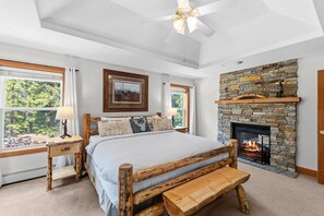 A bedroom with a stone fireplace, a bed with a wooden headboard, a bench, and a window with a view of greenery. - Summit County Escape | Private Hot Tub+ Deck (Breckenridge)