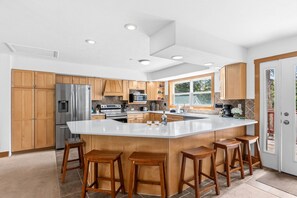 A modern kitchen with wooden cabinets, a large island with stools, stainless steel appliances, and a view to the outside. - Summit County Escape | Private Hot Tub+ Deck (Breckenridge)