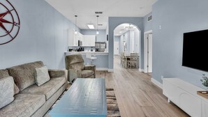 Ein modernes Wohnzimmer mit Sofa, Couchtisch und Fernseher. Es gibt eine Küchenzeile mit Schränken und einem Esstisch.