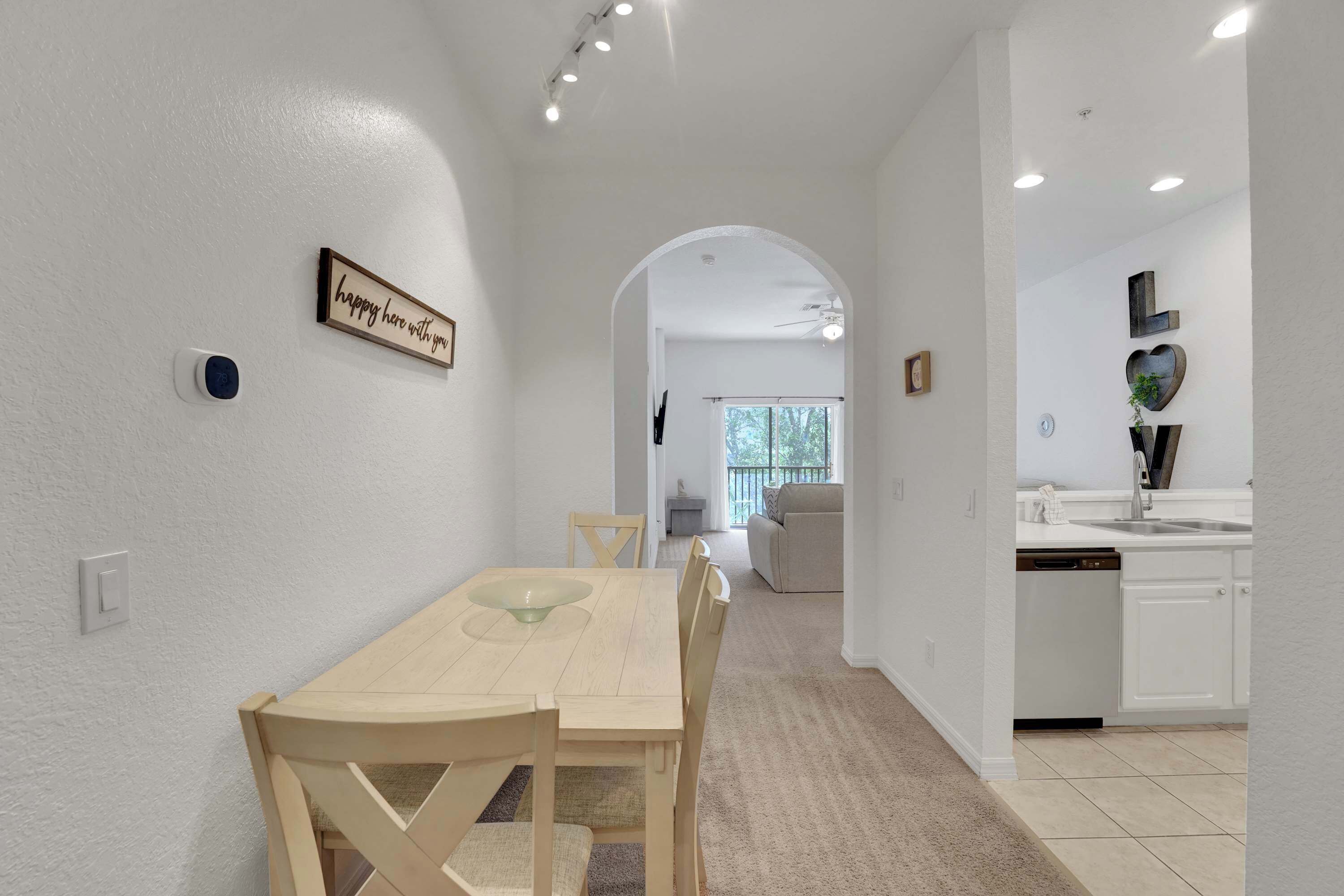 A dining area with a wooden table and chairs, a kitchen with white cabinets, and a hallway leading to a living room.