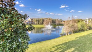 Une vue paisible sur un lac, agrémentée d’une fontaine, entourée de pelouses et d’arbres bien entretenus, avec des bâtiments en arrière-plan.