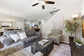 A living room with a gray sofa, patterned armchair, and a coffee table. A ceiling fan is visible, and there is a staircase leading to another level. - Beautiful Home, Themed Rooms, Prime Location (Kissimmee)