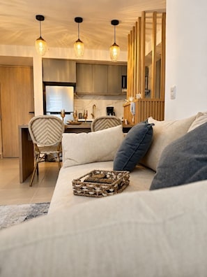 Een moderne woonkamer met een bank, een geweven mand en een eethoek met stoelen, en een keuken op de achtergrond.