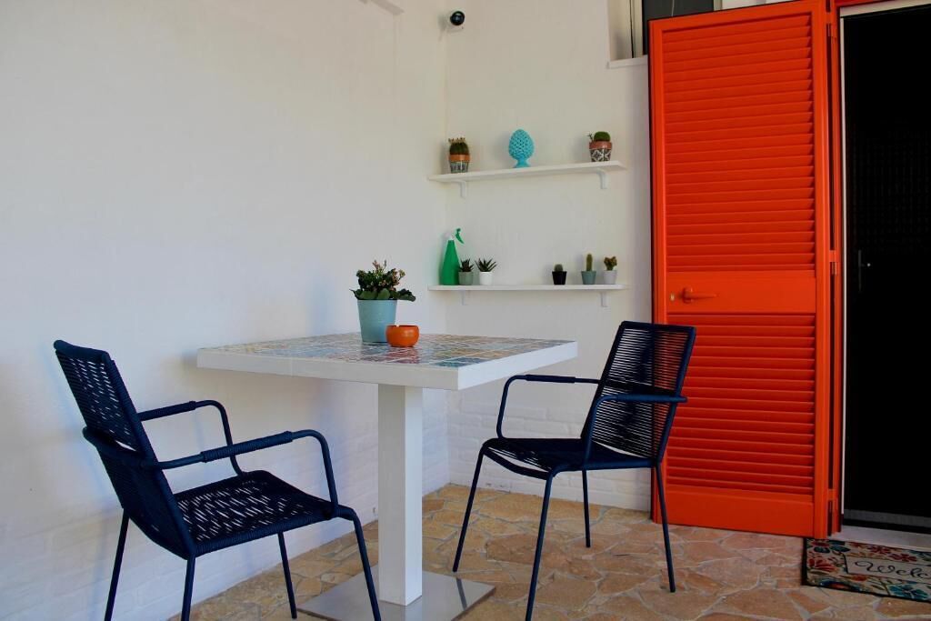 Un petit coin repas avec une table et deux chaises, une porte rouge et des étagères ornées de plantes en pot.