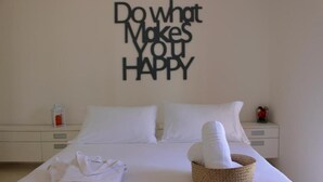 Una cama bien hecha con sábanas blancas, un cartel en la pared que dice 'Haz lo que te haga feliz' y una cesta de toallas sobre la cama.