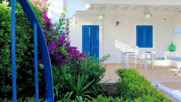 Un patio con mobiliario blanco, contraventanas azules y un jardín con flores moradas.