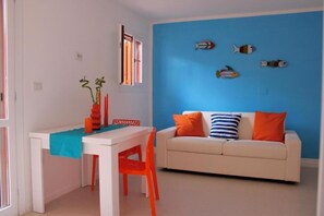 A room with a white table, orange chair, and a white sofa with orange and blue pillows.