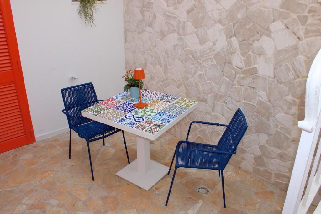 A small outdoor seating area with a tiled table, two blue chairs, a potted plant, and a stone wall.