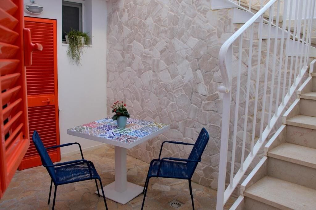 Un mur de pierre, un escalier blanc, une porte rouge et une table avec des chaises.