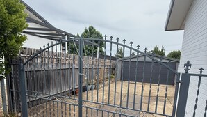 Un espace extérieur clos, délimité par une clôture en métal, avec une clôture en bois en arrière-plan et un bâtiment doté d’un toit en pente.