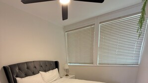 Une chambre à coucher avec un lit, un ventilateur de plafond, une fenêtre avec des stores et une plante.