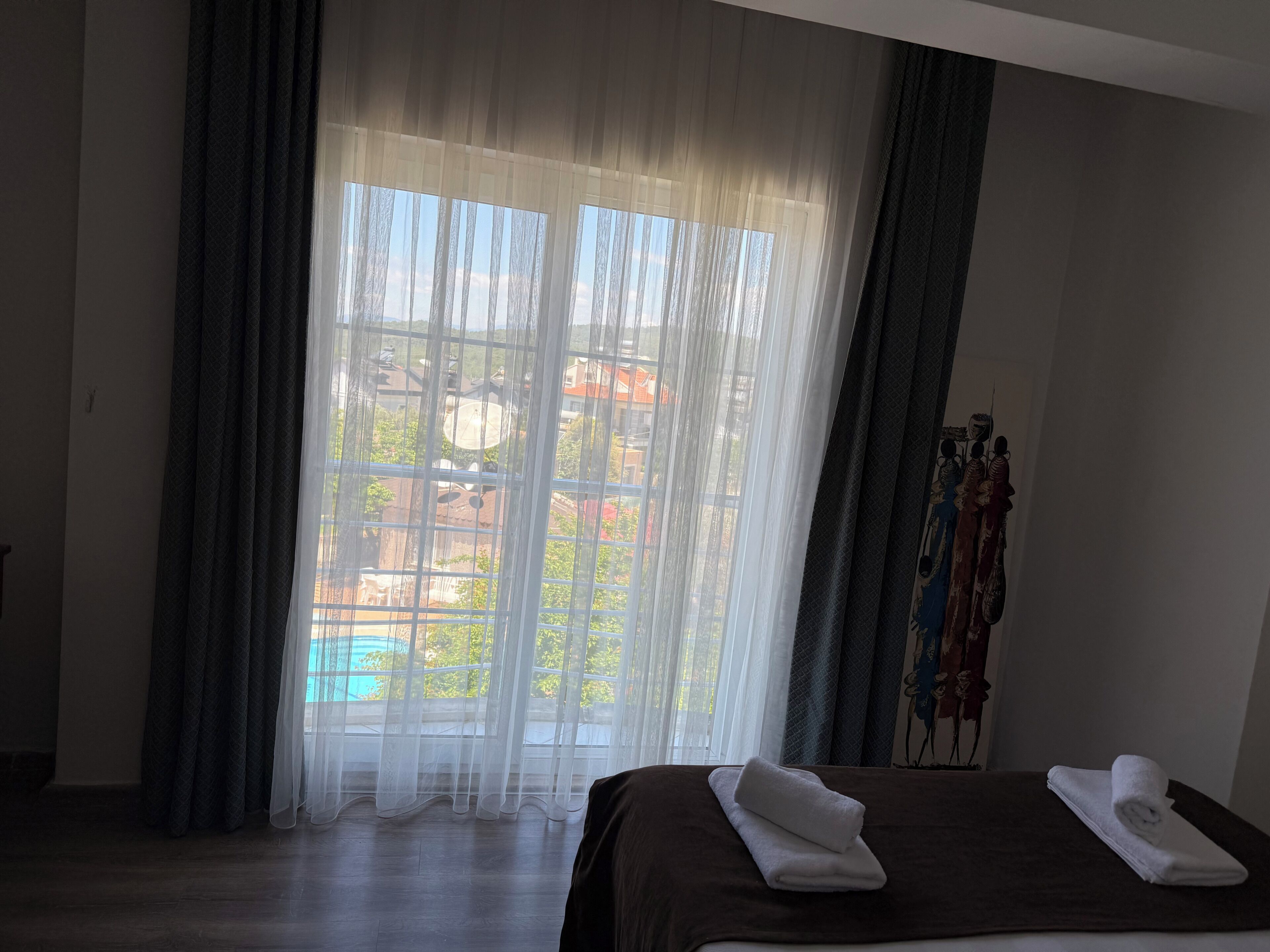 A hotel room with a bed, a window with sheer curtains, and a view of buildings outside.