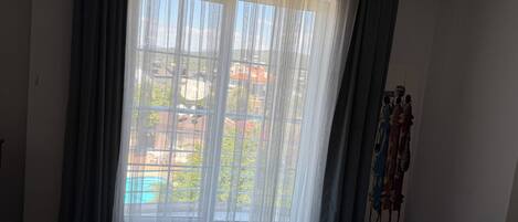 A hotel room with a bed, a window with sheer curtains, and a view of buildings outside.