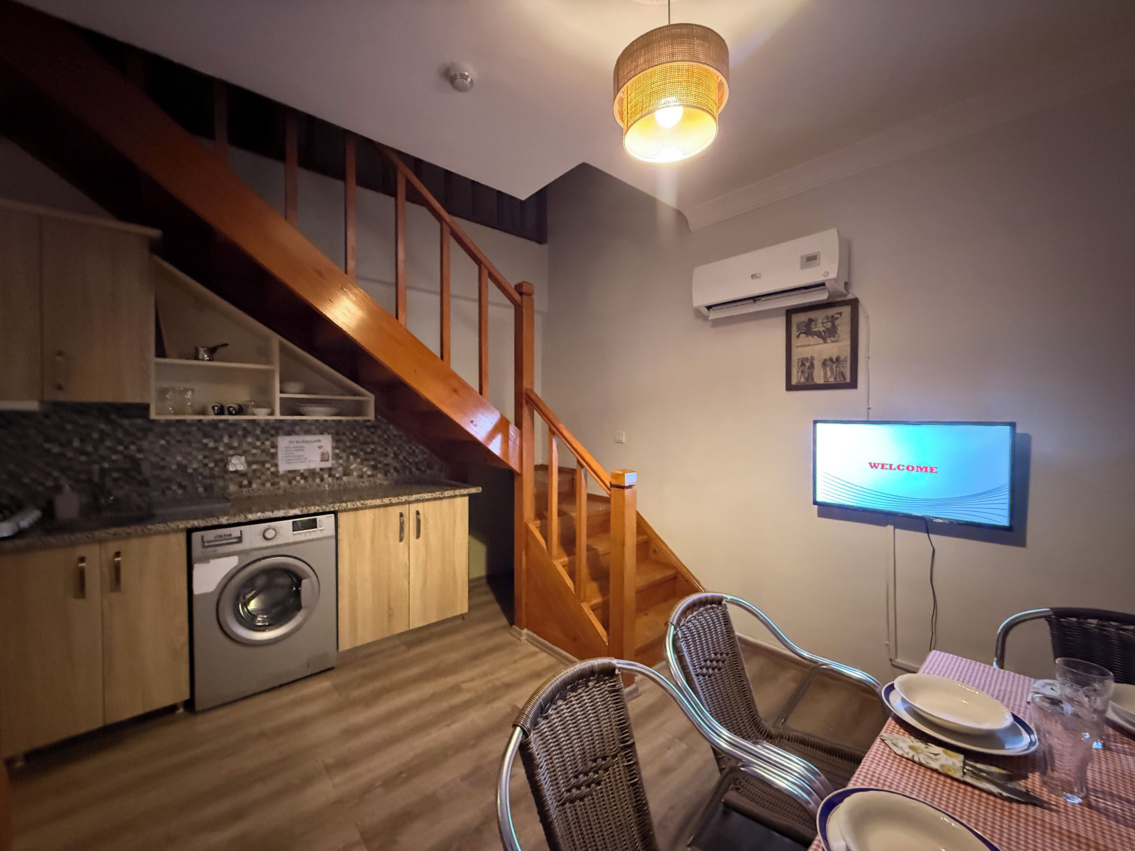 A kitchen with a washing machine, wooden staircase, dining area with chairs, and a TV displaying 'WELCOME'.