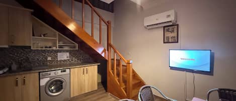 A kitchen with a washing machine, wooden staircase, dining area with chairs, and a TV displaying 'WELCOME'.
