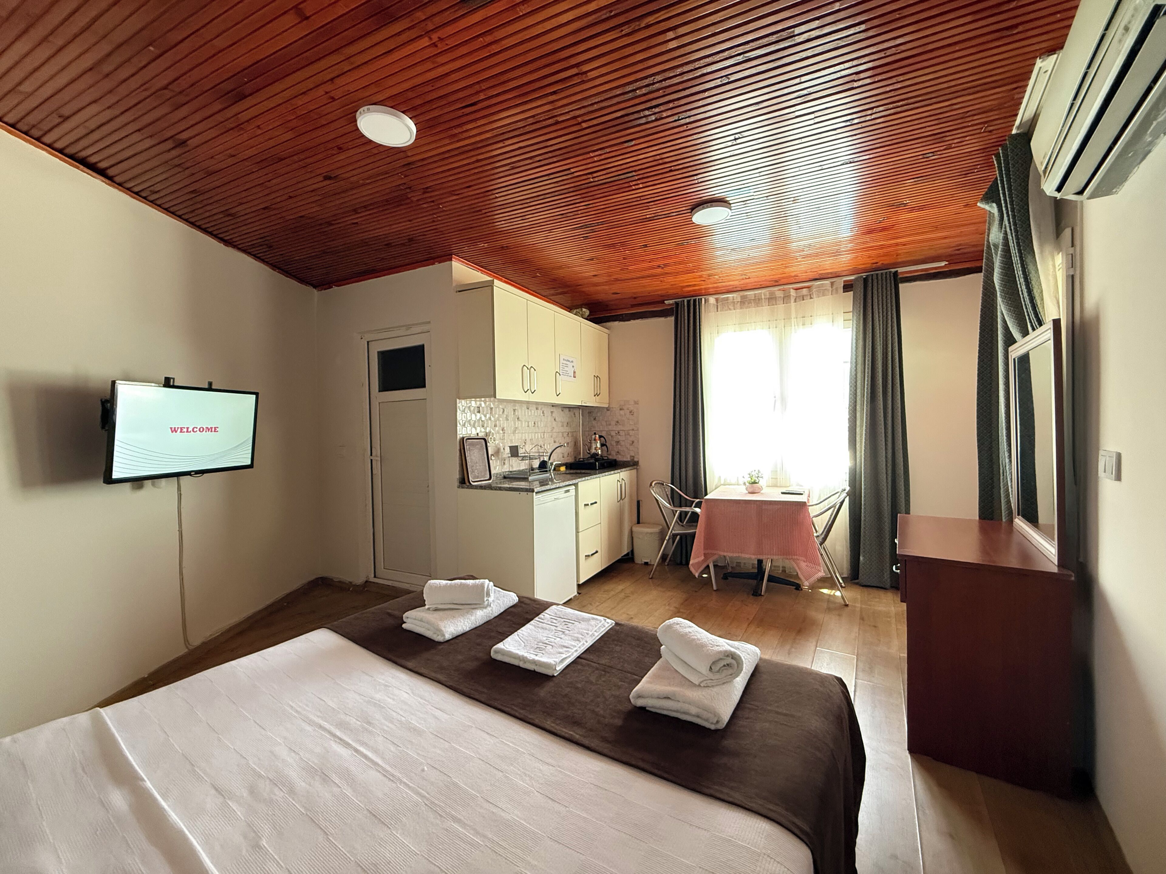 A hotel room with a wooden ceiling, a bed with towels, a small kitchenette, a dining table with chairs, and a TV displaying 'WELCOME'.