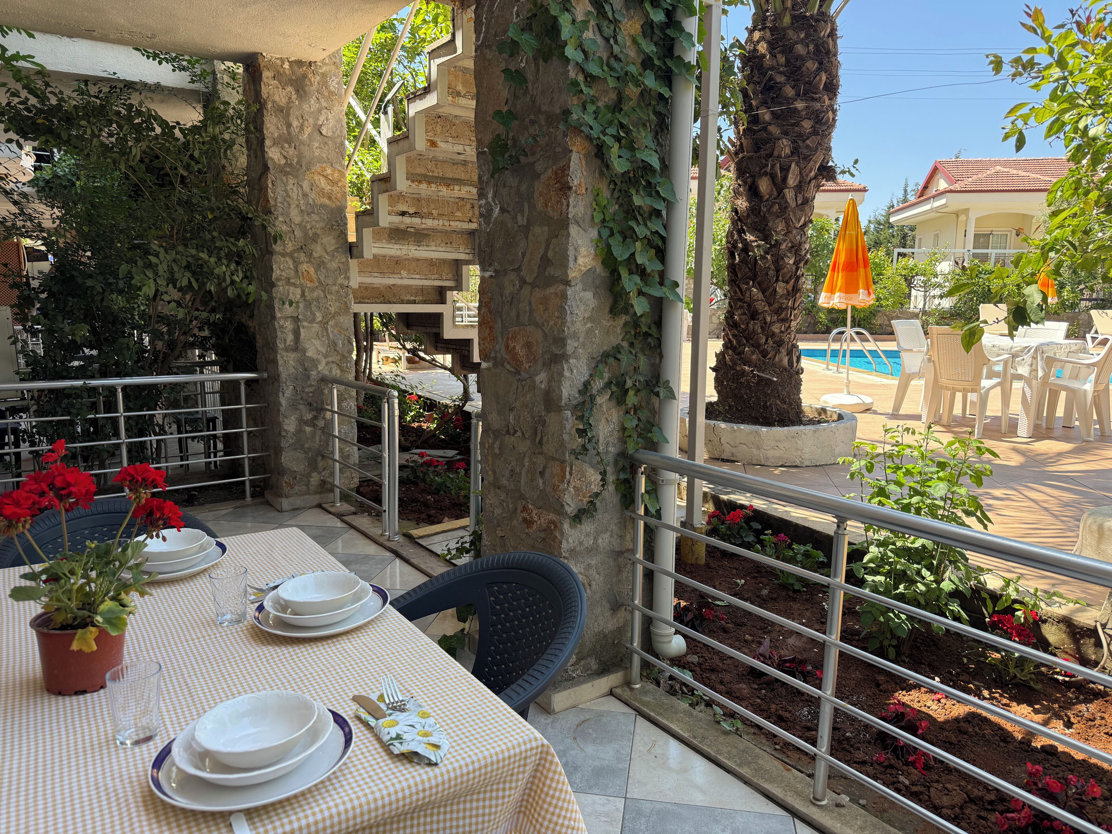 A terrace with a table set for dining, a pool in the background, and a staircase leading to an upper level.