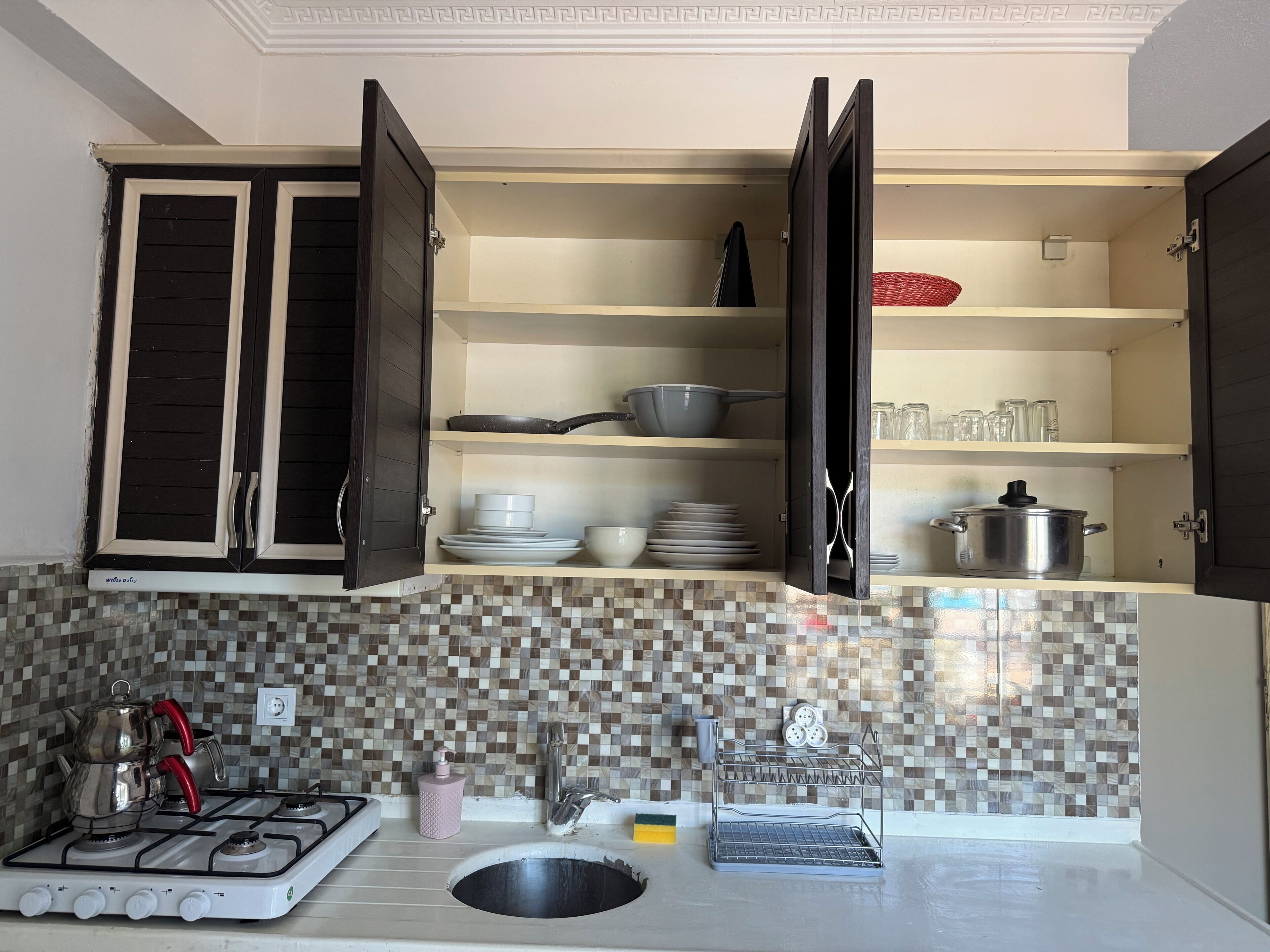 Una cocina con armarios abiertos, estufa a gas, pileta y salpicadero de azulejos.
