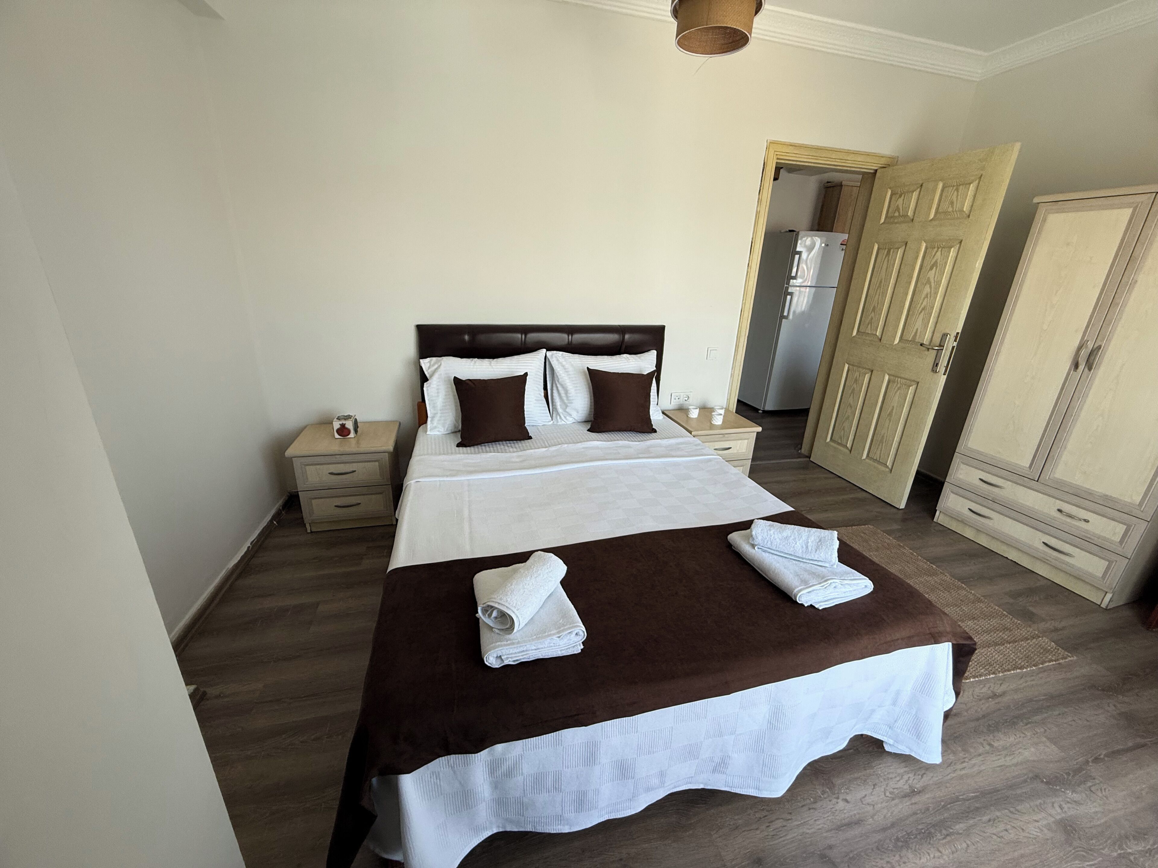 Een net opgemaakt bed met een bruine sprei, witte lakens en twee opgerolde handdoeken. Er staat een houten nachtkastje met een lamp en een klein tafeltje met een kopje en een doosje. Achtergrond: een houten kledingkast.