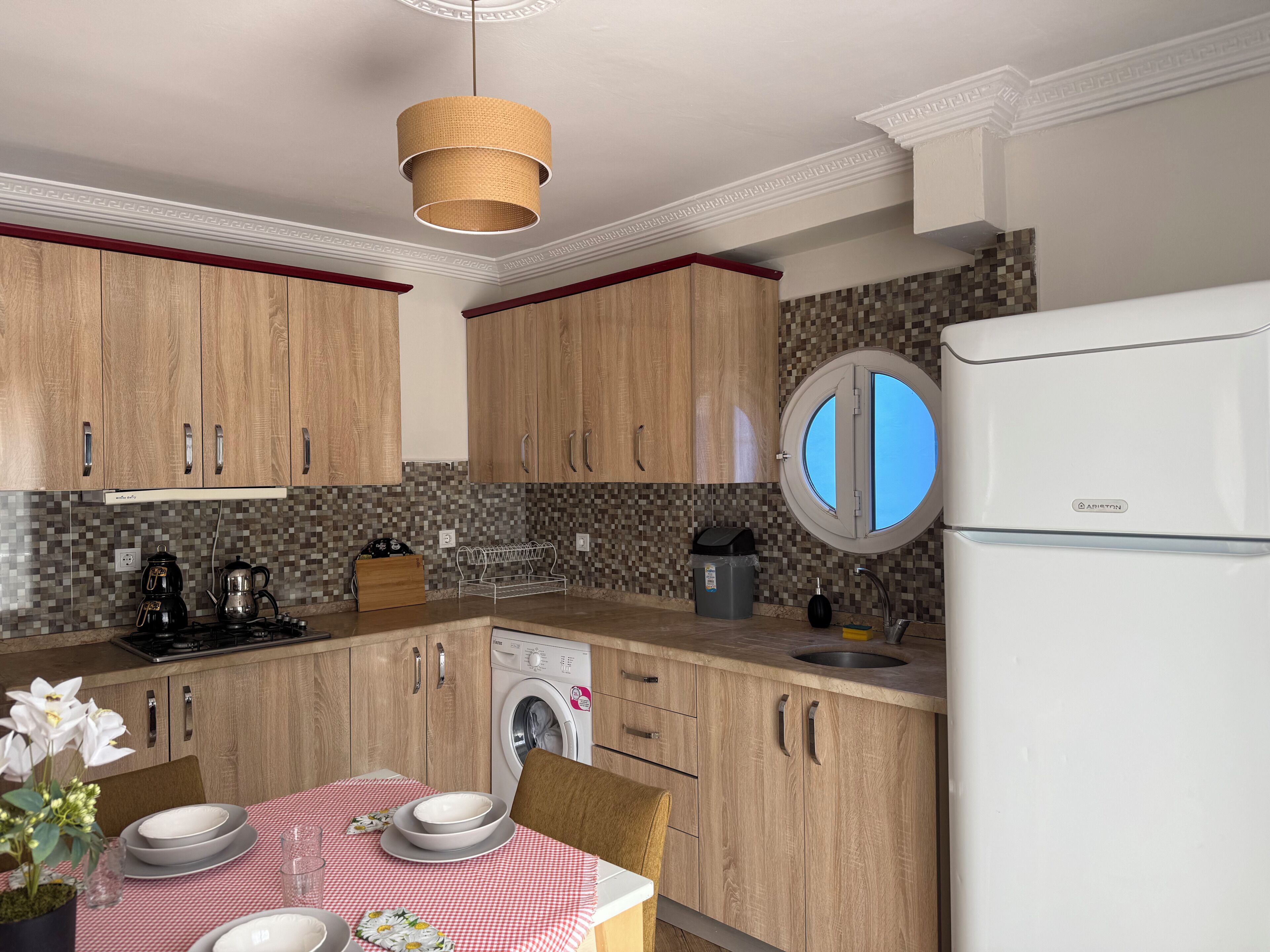 A modern kitchen with wooden cabinets, a round window, a built-in washing machine, and a dining area with a checkered tablecloth.
