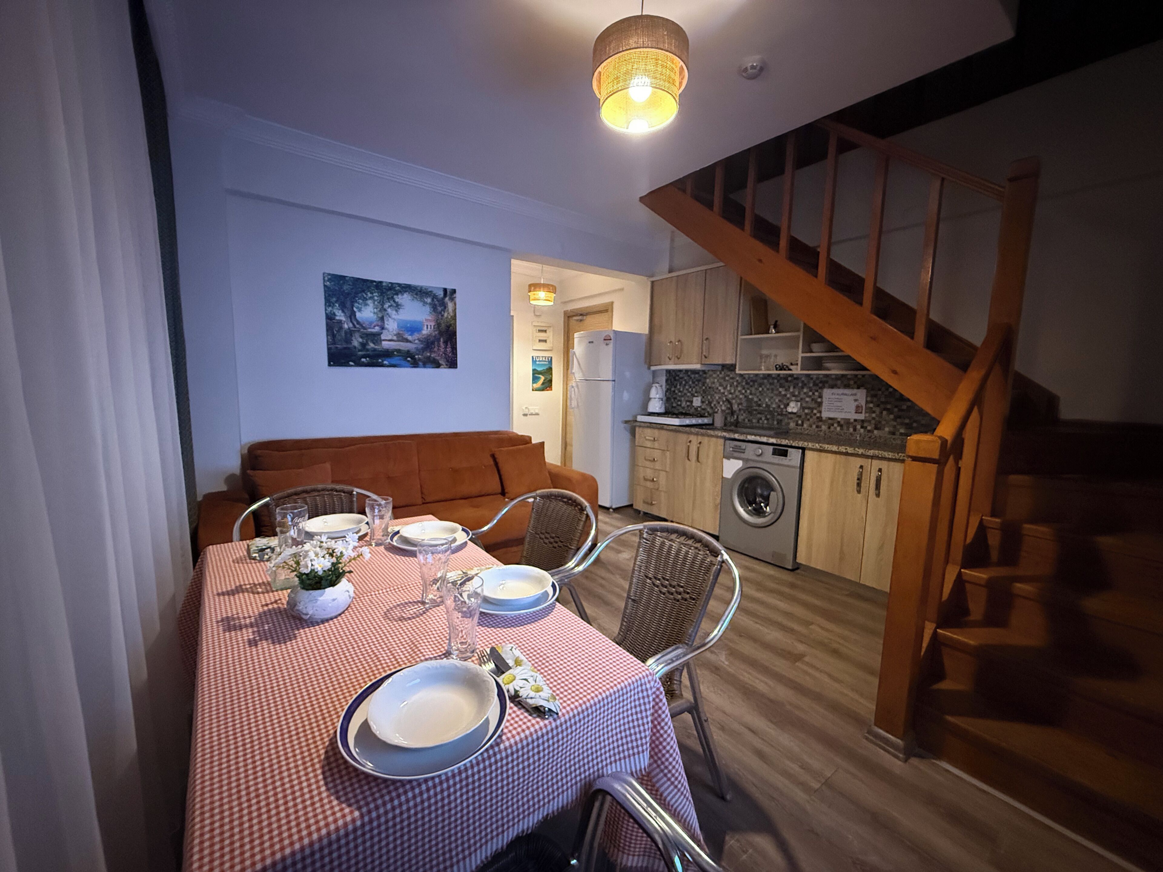 A dining area with a table set for two, a sofa, and a kitchen in the background.