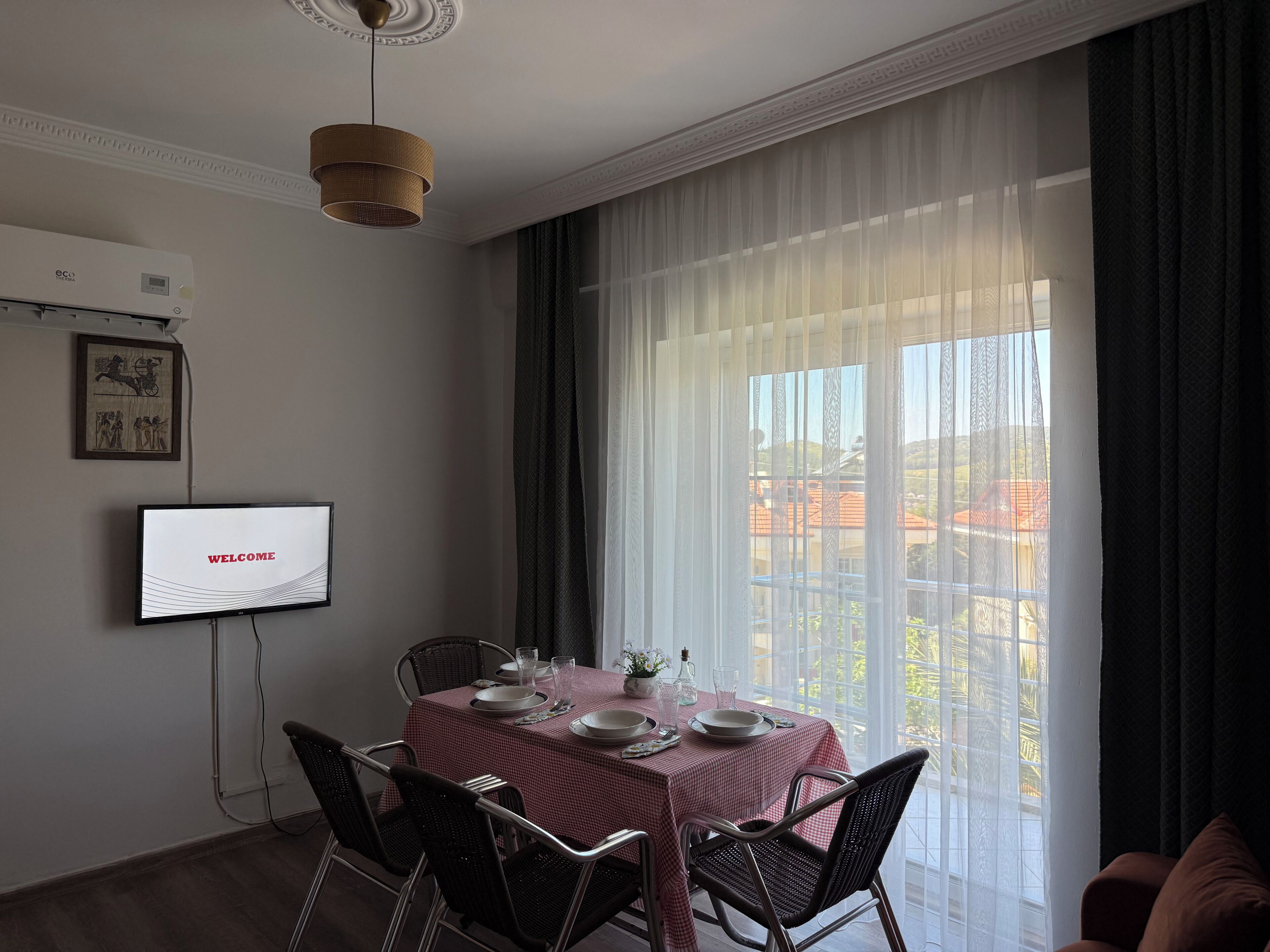 A dining area with a table set for four, a television displaying 'WELCOME', and a large window with sheer curtains.