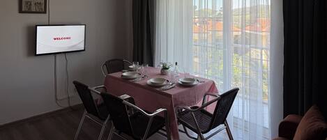 A dining area with a table set for four, a television displaying 'WELCOME', and a large window with sheer curtains.