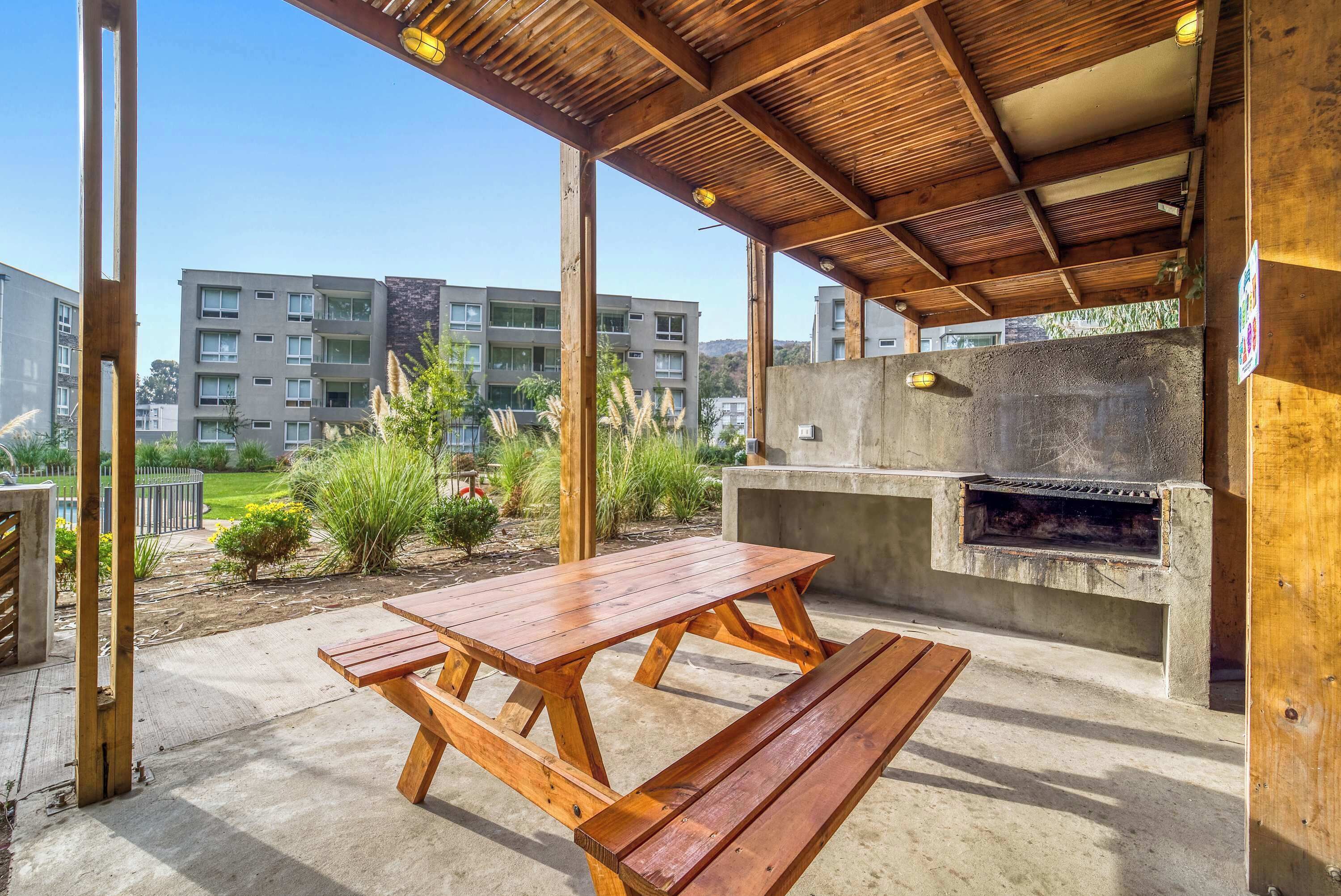Eine Terrasse im Freien mit einem Holztisch und Bänken, einem Grillbereich aus Beton und Blick auf Wohnhäuser im Hintergrund.