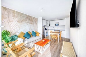 A modern living room with a sofa, coffee table, and chairs, a kitchen with white cabinets, and a dining area with a table and chairs.
