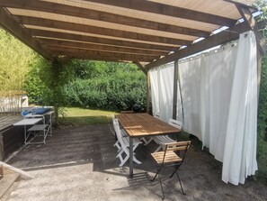 Un patio couvert avec une table et des chaises en bois, un barbecue et un rideau blanc.