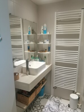 Une salle de bain avec un lavabo blanc, un miroir et des étagères contenant des articles de toilette. On y trouve également des toilettes et une poubelle.