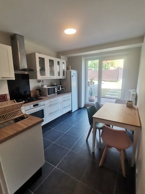 A modern kitchen with white cabinets, a stainless steel range hood, a microwave, and a refrigerator. There is a dining table with chairs and a view of a backyard through a sliding glass door. - A quiet, self-contained home surrounded by greenery (Villersexel)
