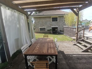 Une terrasse couverte avec une table et des chaises en bois, un bâtiment en pierre en arrière-plan et une cour clôturée.