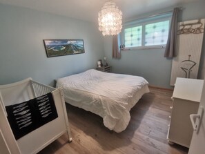 A bedroom with a bed, a crib, a chandelier, a window with curtains, a radiator, and a wall-mounted shelf with decorative items. - A quiet, self-contained home surrounded by greenery (Villersexel)
