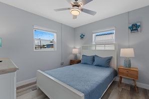 Un dormitorio con cama, mesitas de noche, ventilador de techo y dos ventanas.