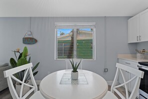 Una cocina moderna con muebles blancos, una mesa de comedor con sillas, una planta en maceta y vistas a un paisaje verde a través de una ventana.